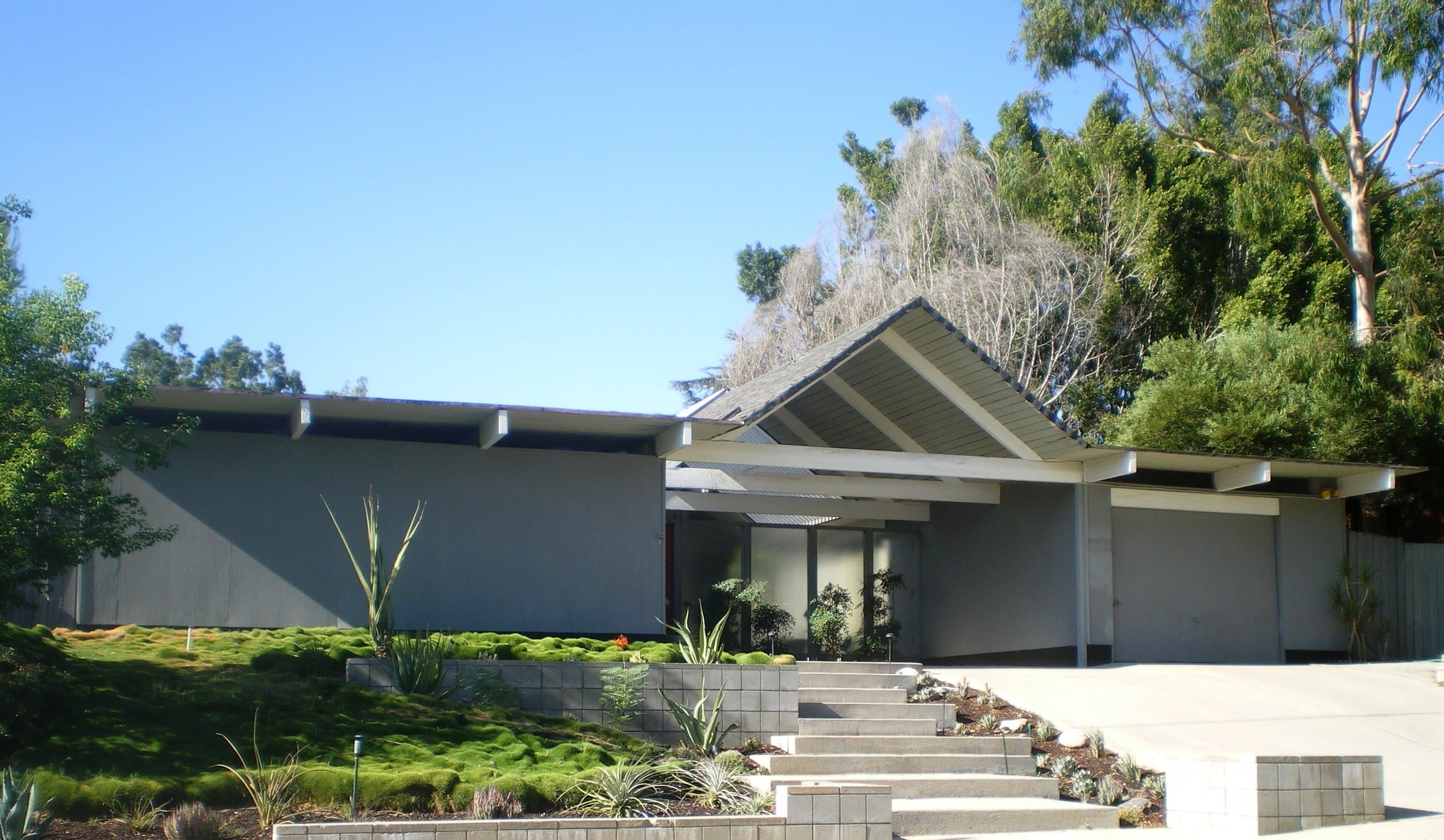 An Eichler with an open gable on exposed beams sheltering a glass-walled entry, above terraced front steps