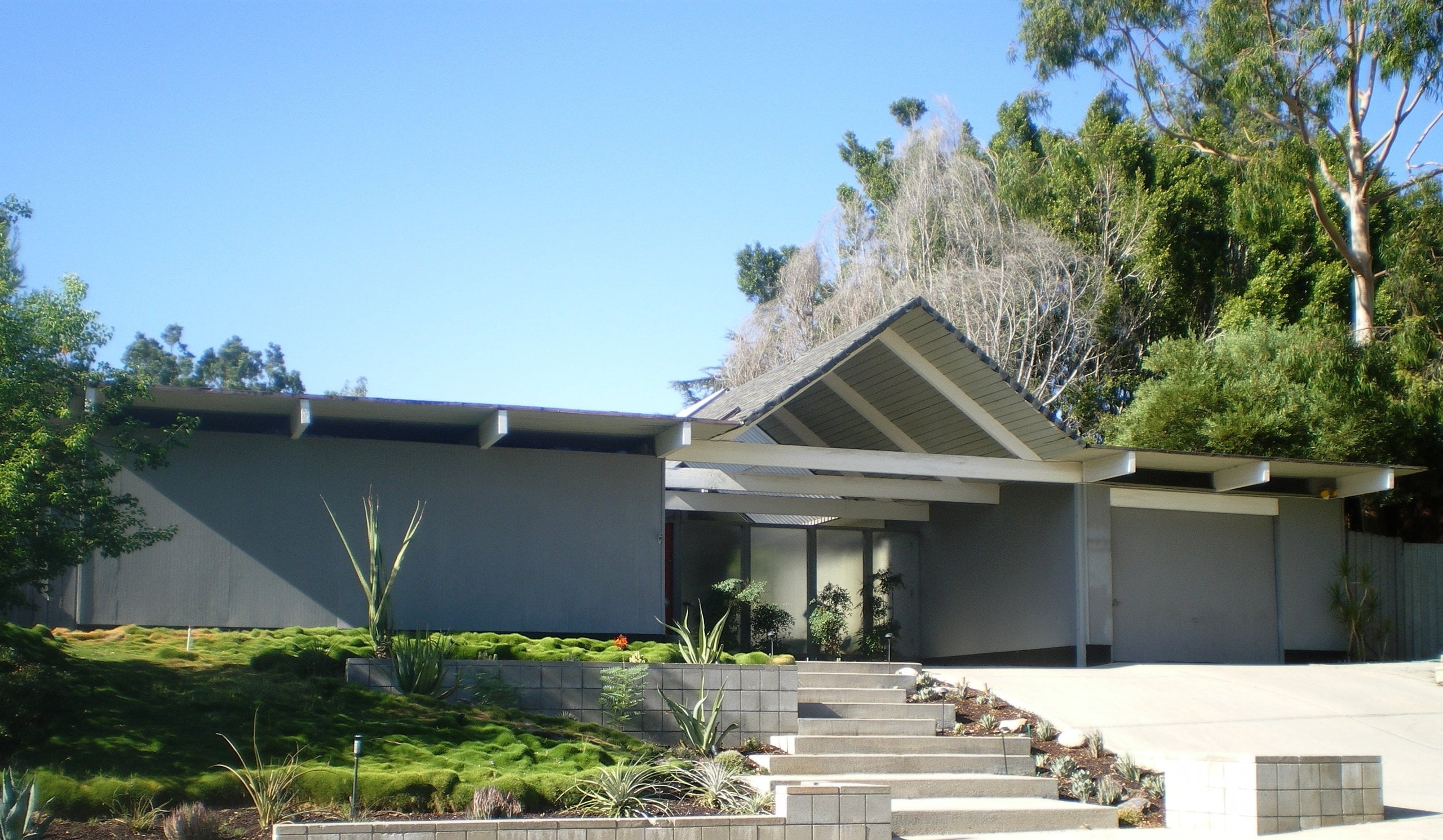 An Eichler with an open gable on exposed beams sheltering a glass-walled entry, above terraced front steps