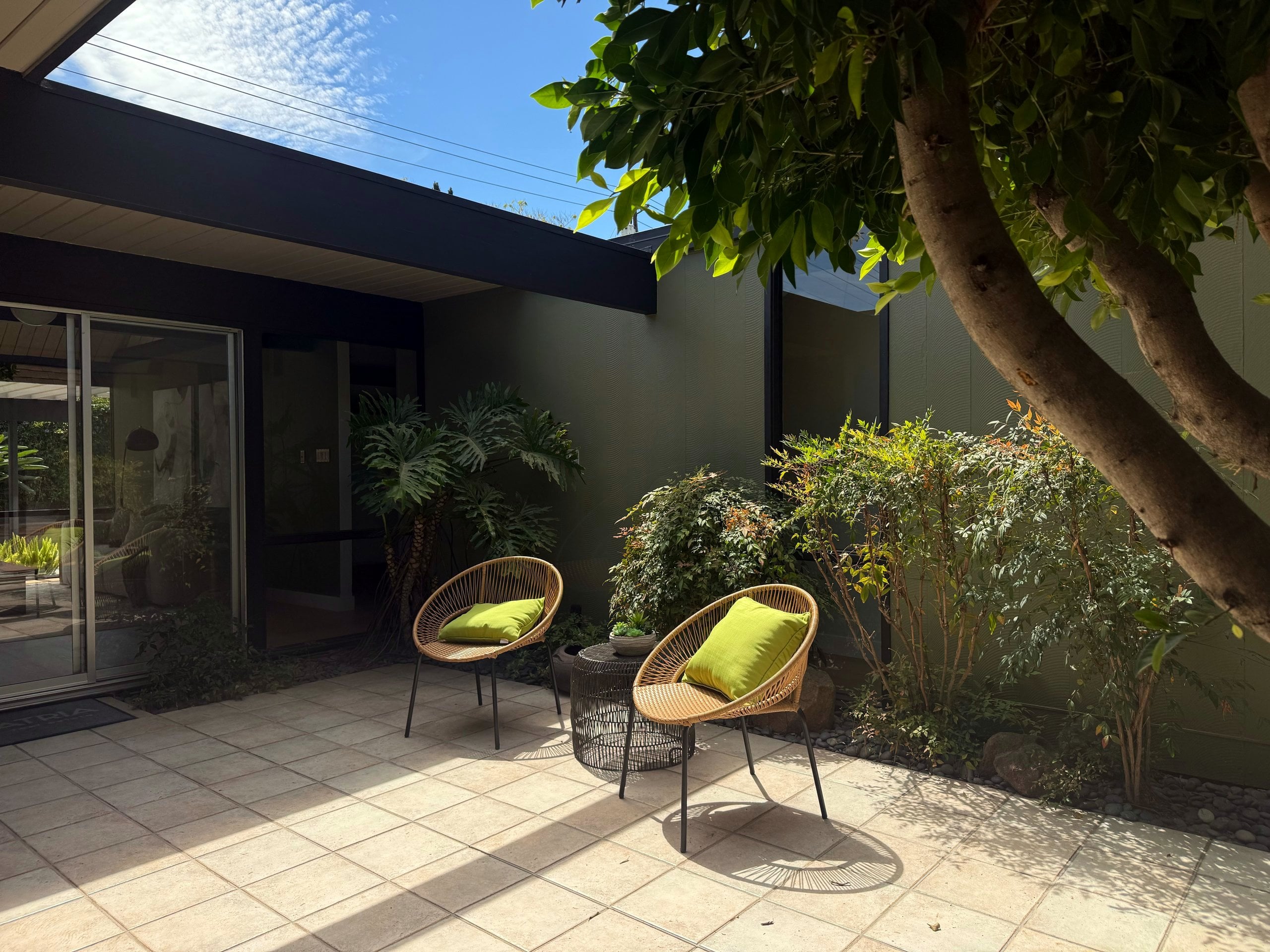 An open-air atrium patio with two rattan chairs, framed by glass walls and a tree