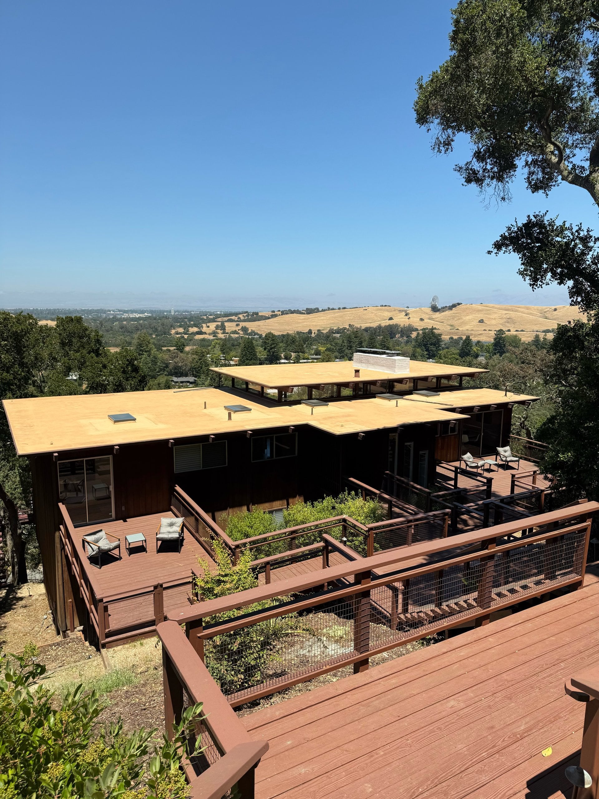 A mid-century home with flat roofs and wraparound wood decks stepping down a wooded hillside above golden hills