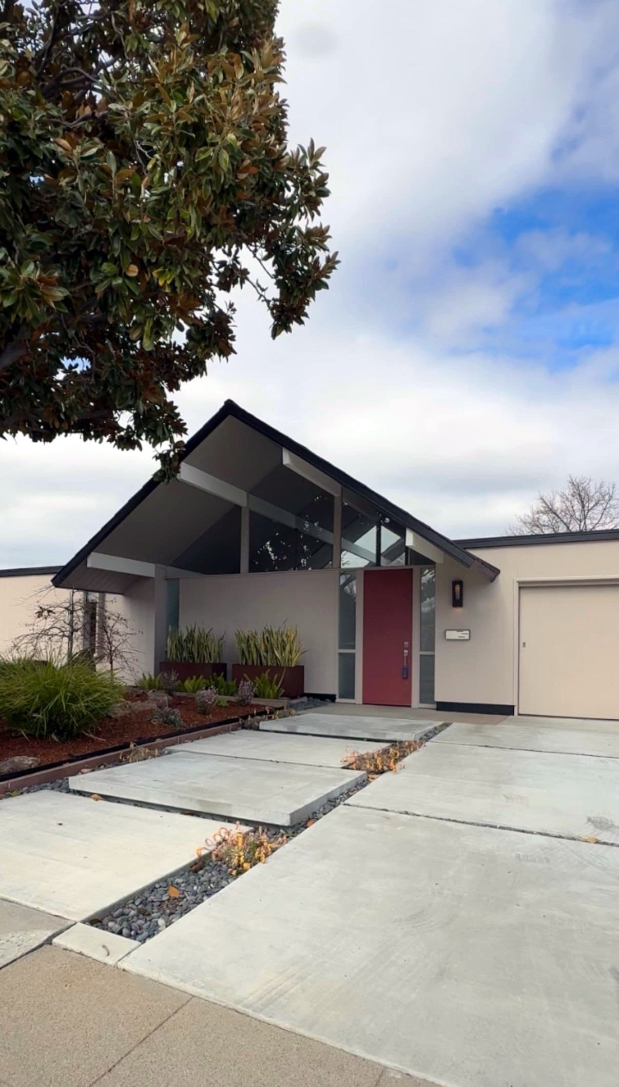 An Eichler with an open gable over clerestory glass and a red front door, behind a walkway of offset concrete pads