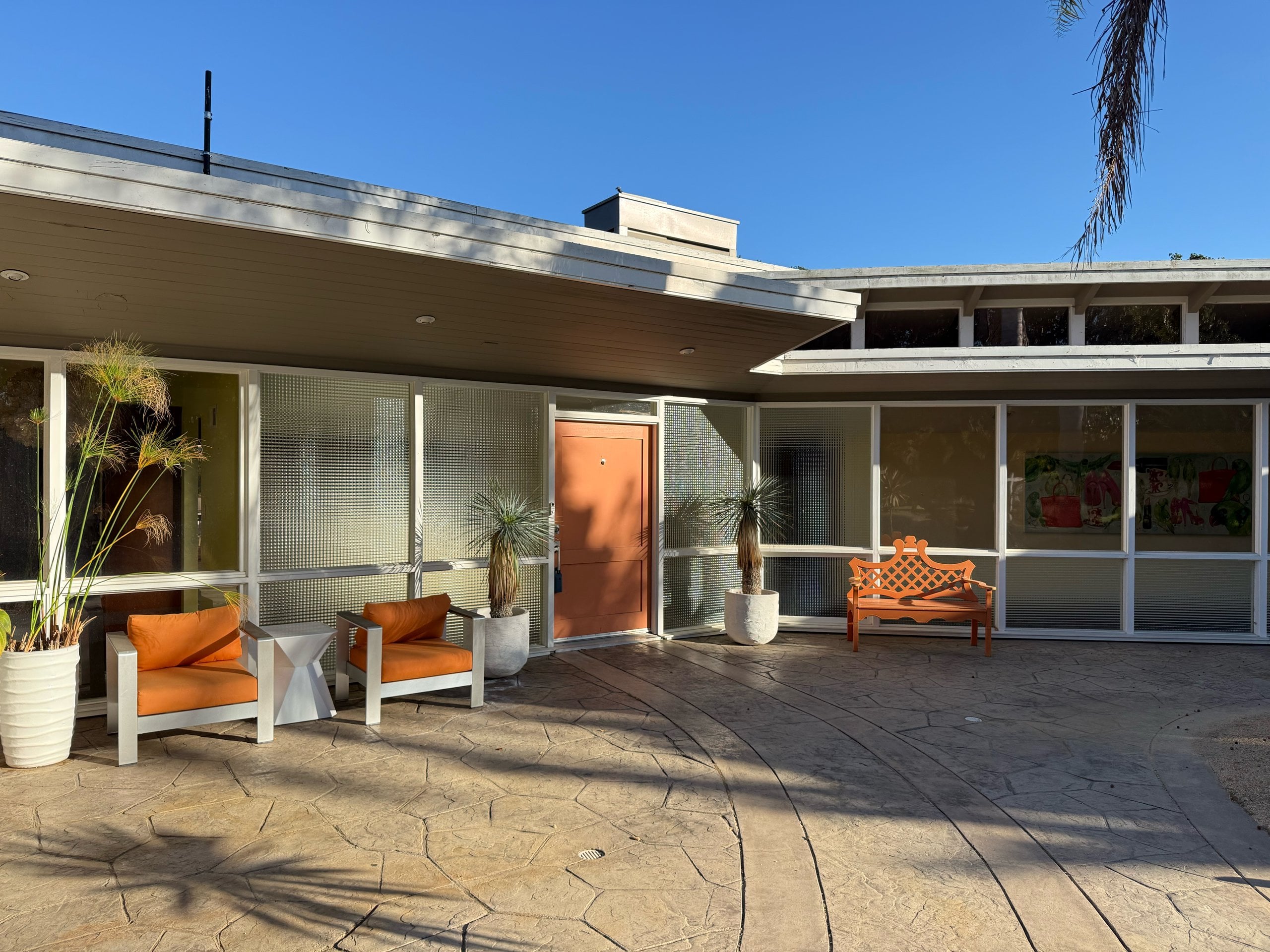 A mid-century courtyard facade of glass panels under a deep overhang, with an orange front door