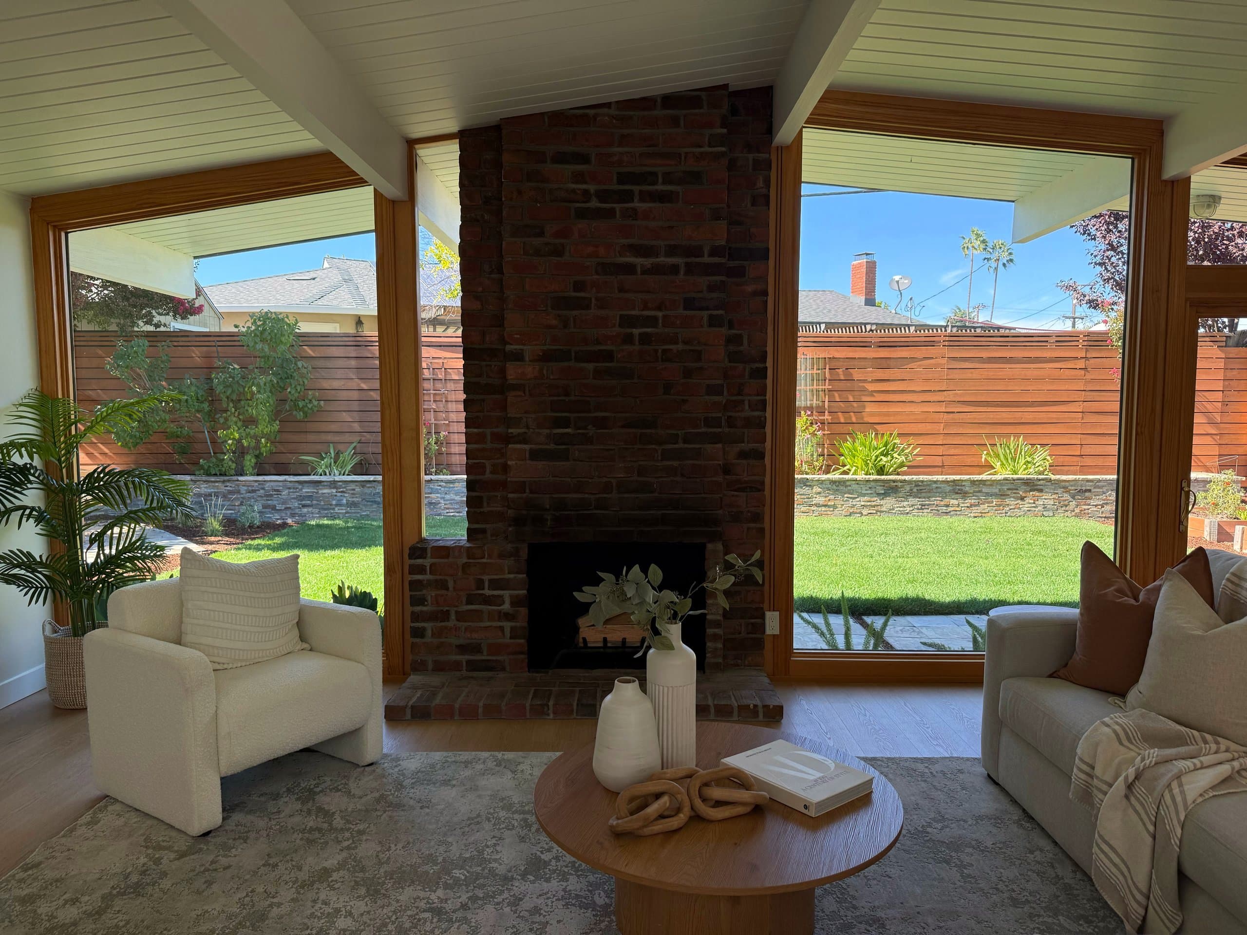 A living room with a brick fireplace between floor-to-ceiling windows looking onto a green backyard