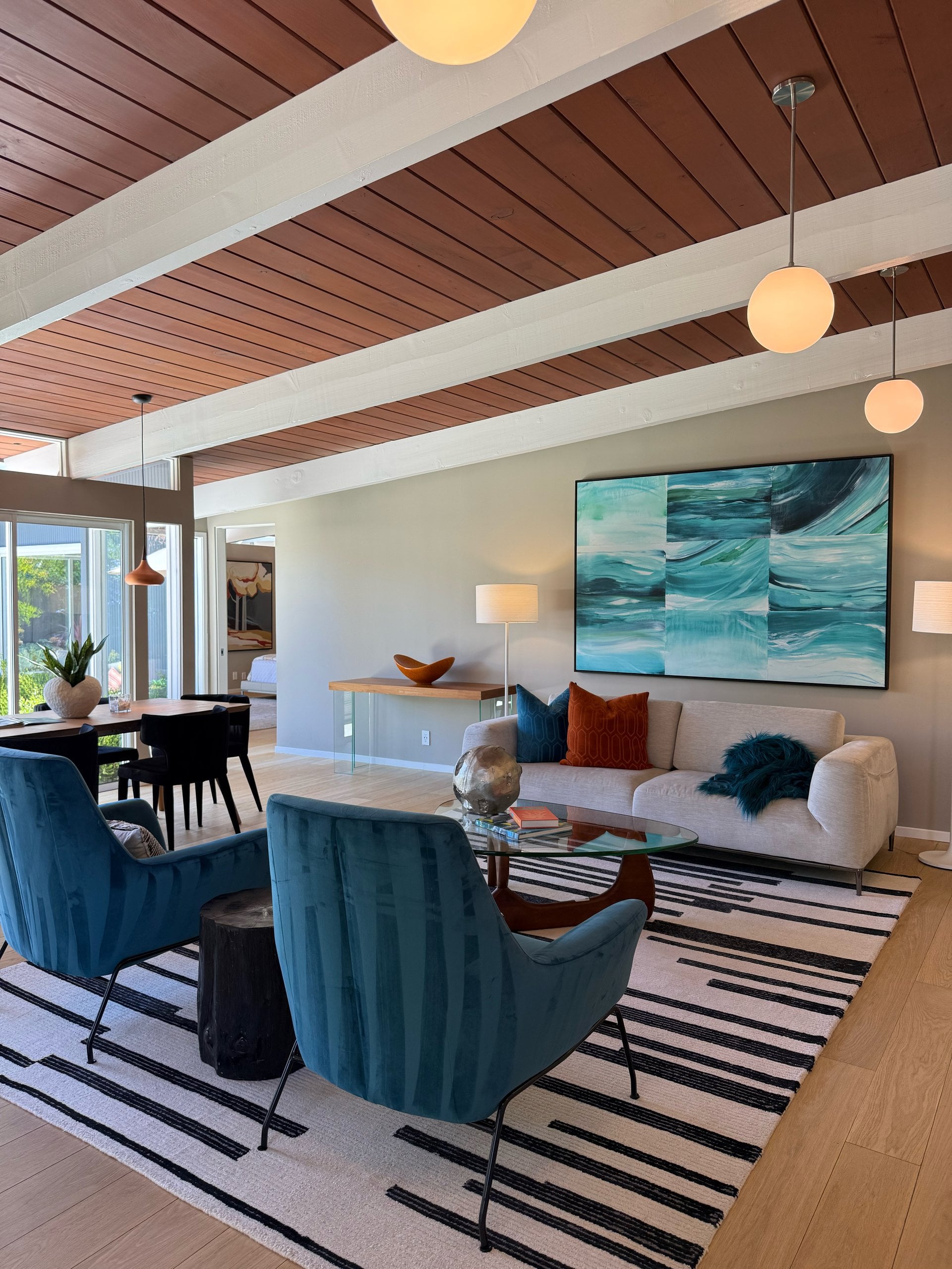 A staged living room under a wood ceiling, with teal velvet chairs, a cream sofa, and a large blue painting